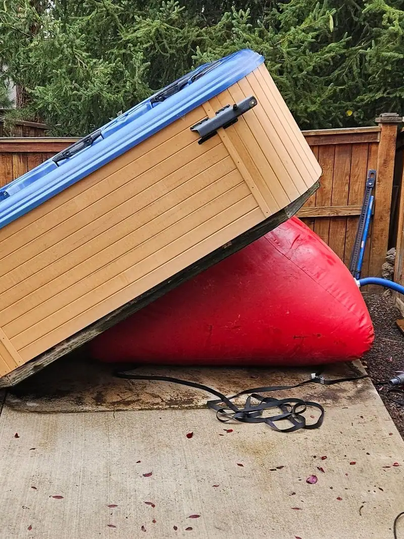 An old hot tub tipped up on a red inflatable lift beside a trailer on a deck